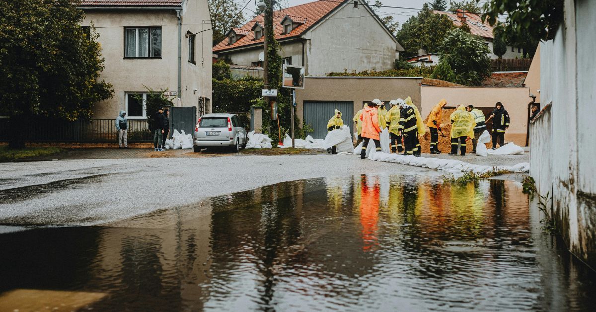 Emergency responders and local volunteers in protective gear place sandbags around homes to prevent further flood damage in a residential neighborhood. Floodwaters have risen onto the street, reflecting the overcast sky and rescue efforts.