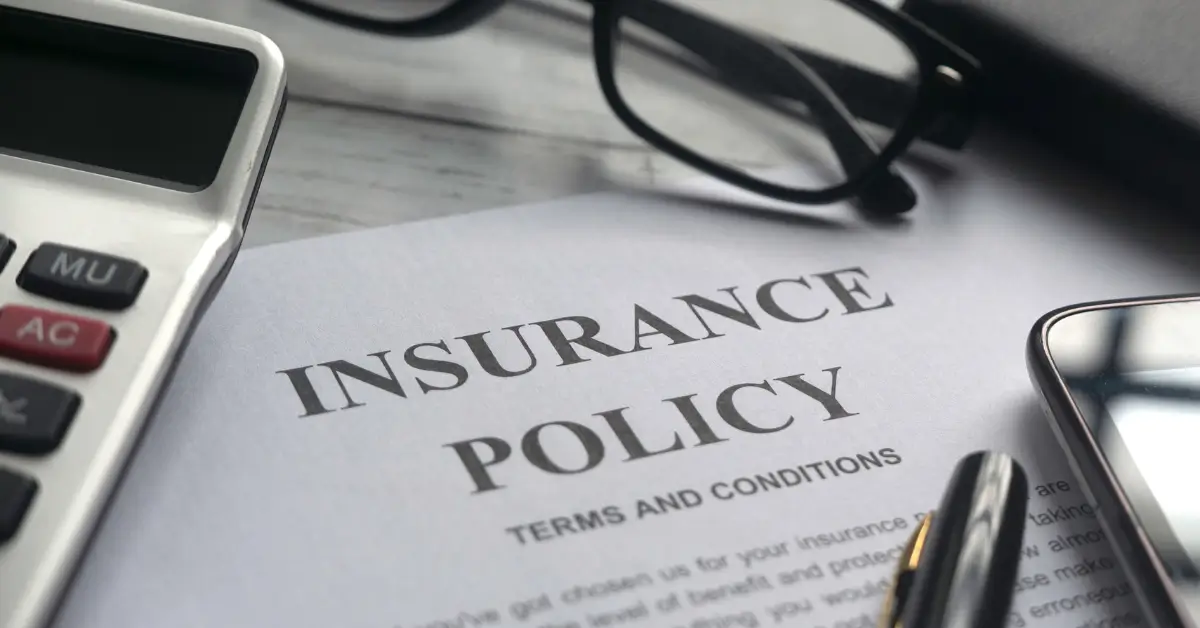 Close-up of an insurance policy document with the words 'INSURANCE POLICY' prominently visible, surrounded by a pair of glasses, a pen, a calculator, and a smartphone on a desk.