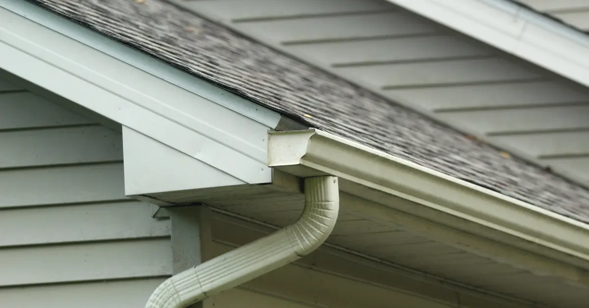 Close-up view of a house’s roofline showing clean gutters and a downspout, illustrating proper water drainage maintenance to prevent basement flooding.