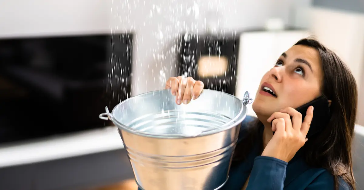Woman holding a metal bucket under a leaking ceiling to catch dripping water while talking on the phone, looking concerned.