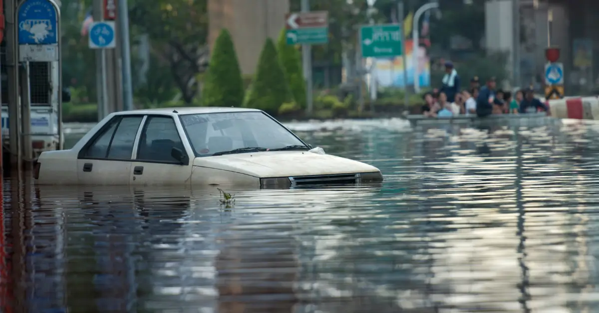White car partially submerged in floodwater on a city street, with people and emergency personnel visible in the background."